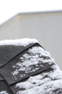 Roofing material samples beside a house with asphalt shingles and a metal roof during a snowy winter.