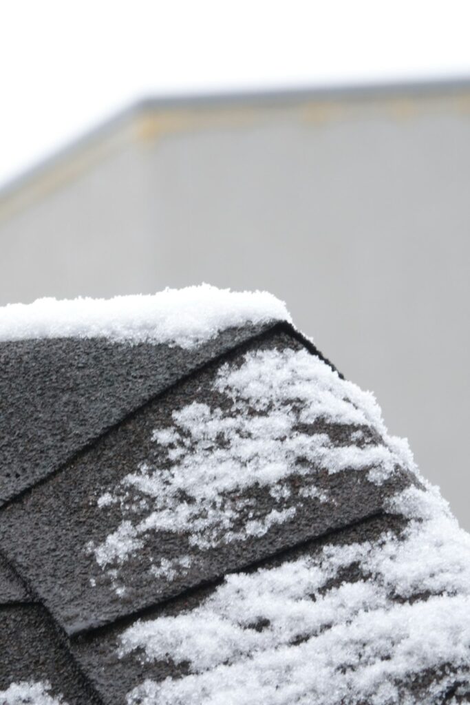 Roofing material samples beside a house with asphalt shingles and a metal roof during a snowy winter.